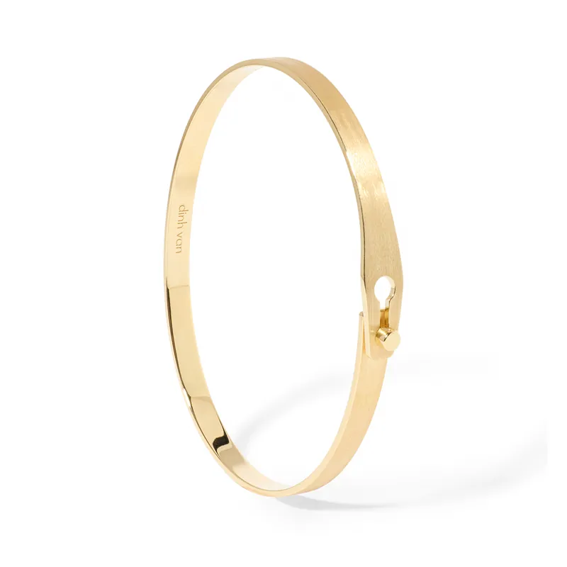 Serrure bangle in brushed yellow gold: a slim open band whose flattened end forms a keyhole-shaped clasp, on a white ground.