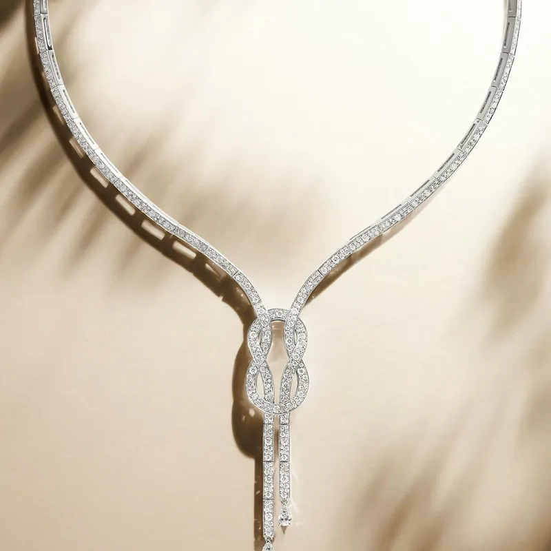 Chance Infinie necklace in white gold paved with diamonds, a figure-eight knot at the front with two drops, on a sunlit beige ground