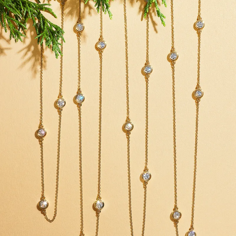 Elsa Peretti Diamonds by the Yard sprinkle necklaces in 18k yellow gold, bezel-set diamonds spaced along fine chains, hanging against a peach ground with evergreen sprigs.