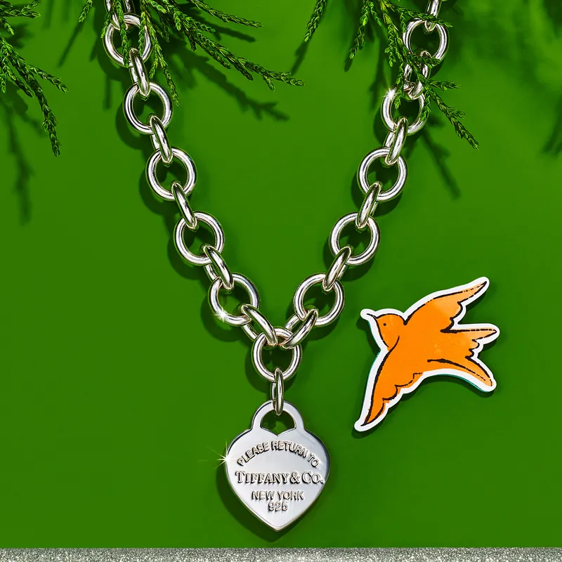 Return to Tiffany heart tag chain link necklace in sterling silver, the tag engraved Please Return to Tiffany & Co. New York, on a green ground beside an orange Andy Warhol bird from the 2022 holiday campaign.