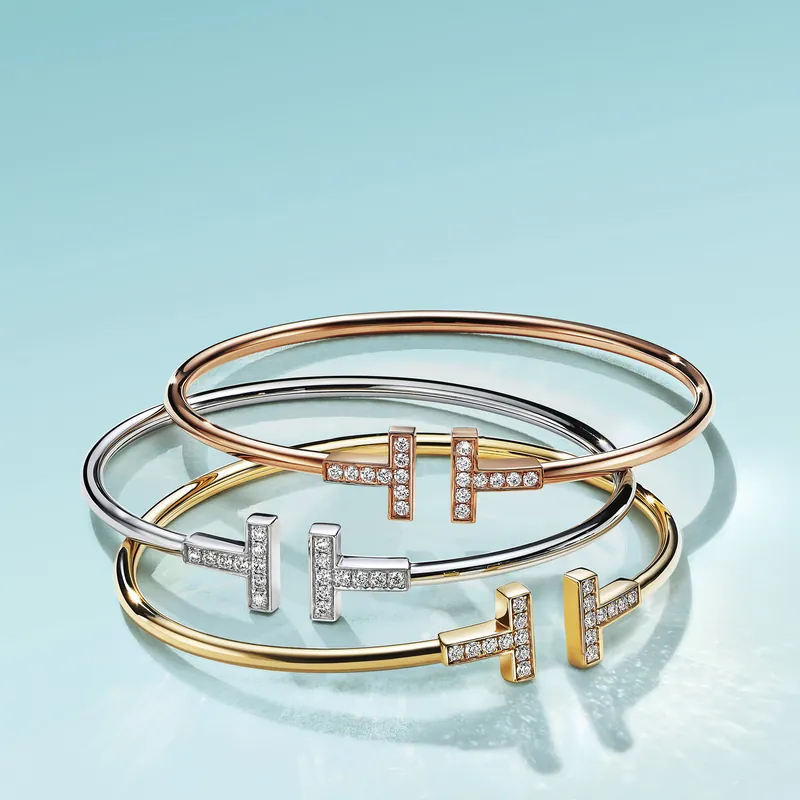 Three Tiffany T wire bracelets in rose, white and yellow gold, their T-shaped terminals set with diamonds, stacked on a pale blue ground.